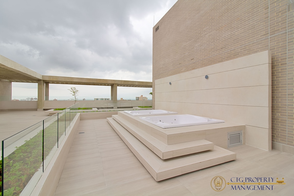 An outdoor terrace features a white jacuzzi set on a series of steps, surrounded by sleek design elements. Large glass panels provide a clear view of the surrounding scenery while allowing natural light to enhance the area.