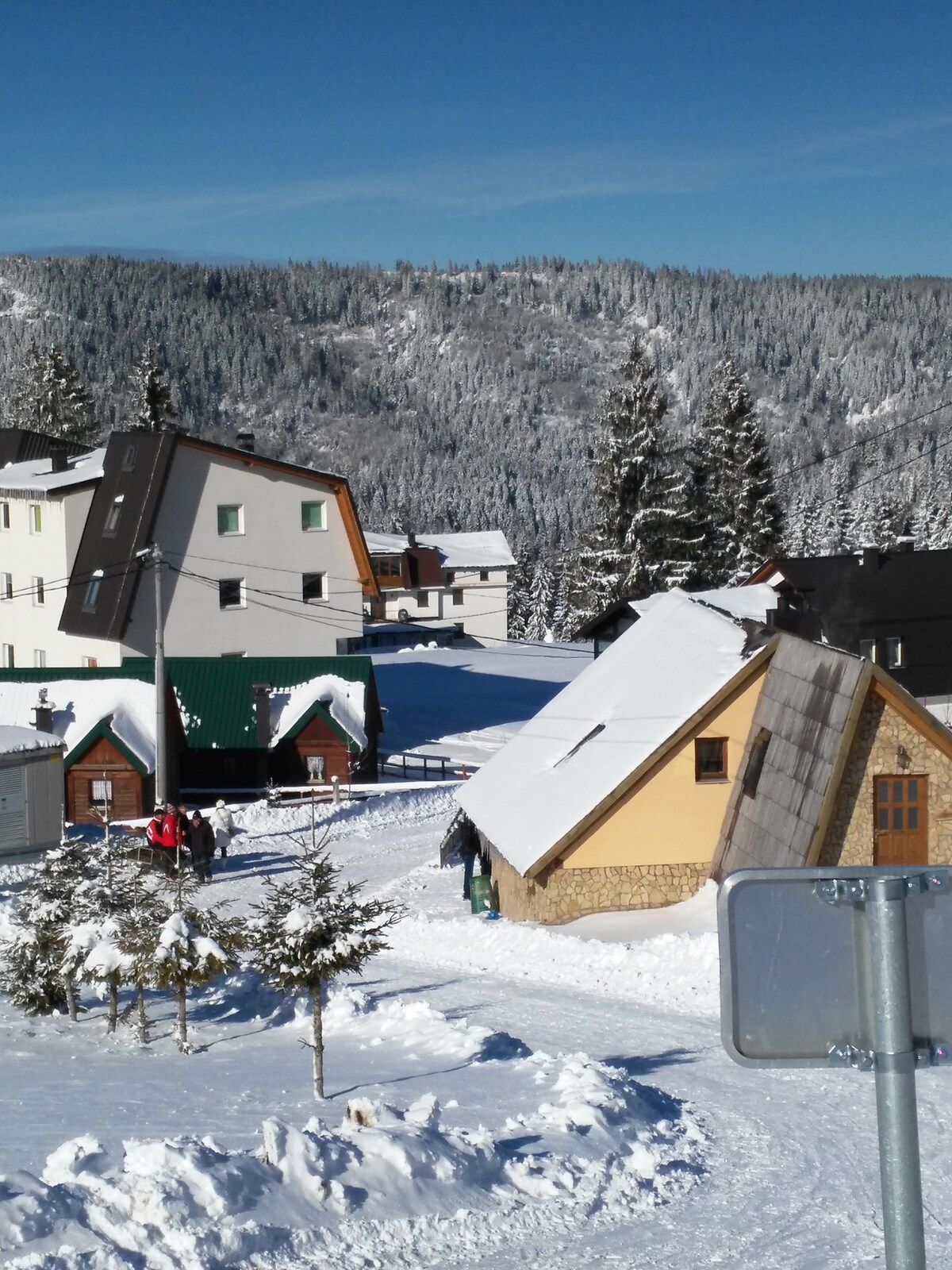 A winter scene displays a village surrounded by snow-covered hills, with wooden and modern structures nestled among evergreen trees. A couple of people can be seen walking along the snow-covered path, while the clear blue sky provides a bright backdrop.