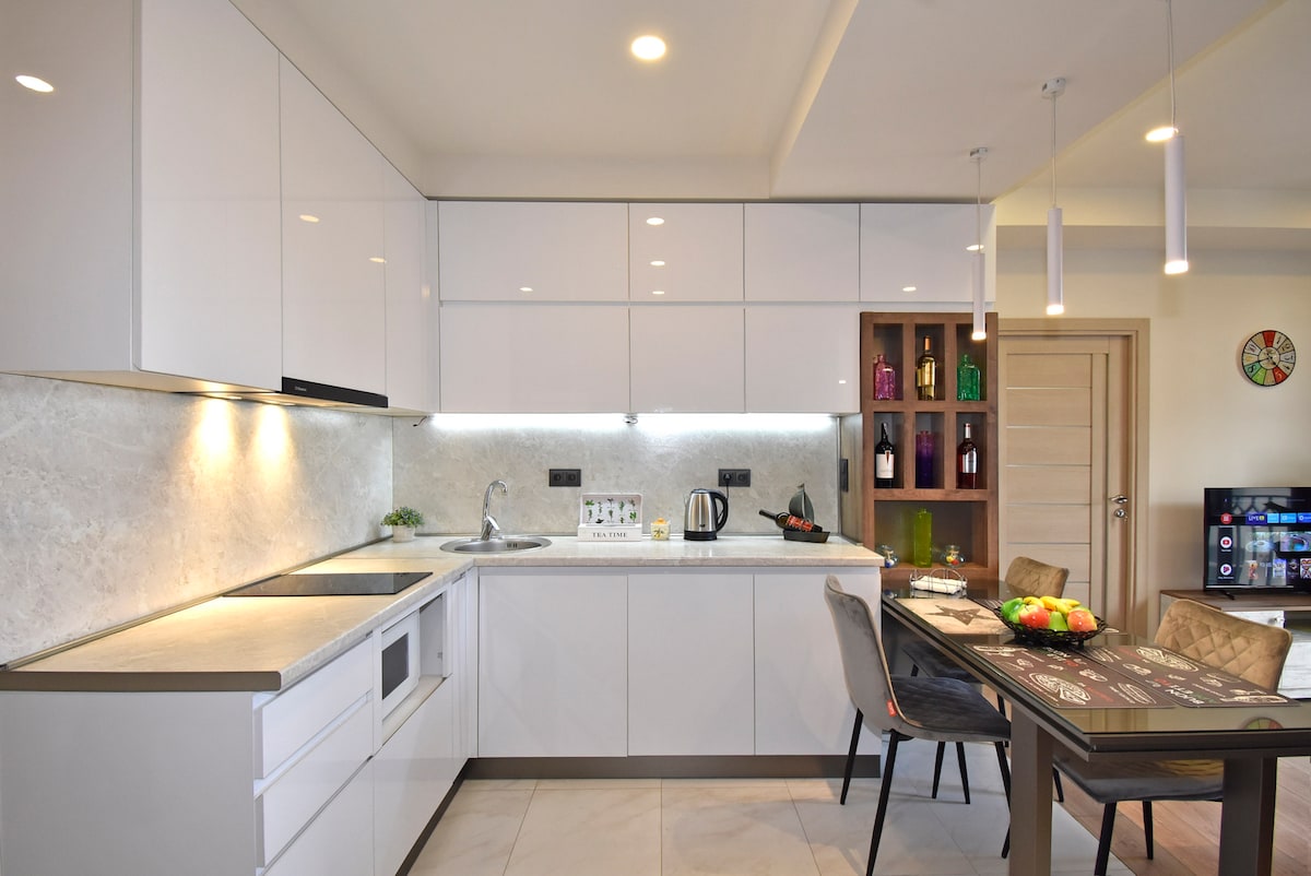 The kitchen area features sleek white cabinetry and a modern countertop. A variety of cooking appliances, including a kettle and microwave, are present. A table is set with fresh fruit, and three dining chairs are arranged around it, enhancing the inviting space.