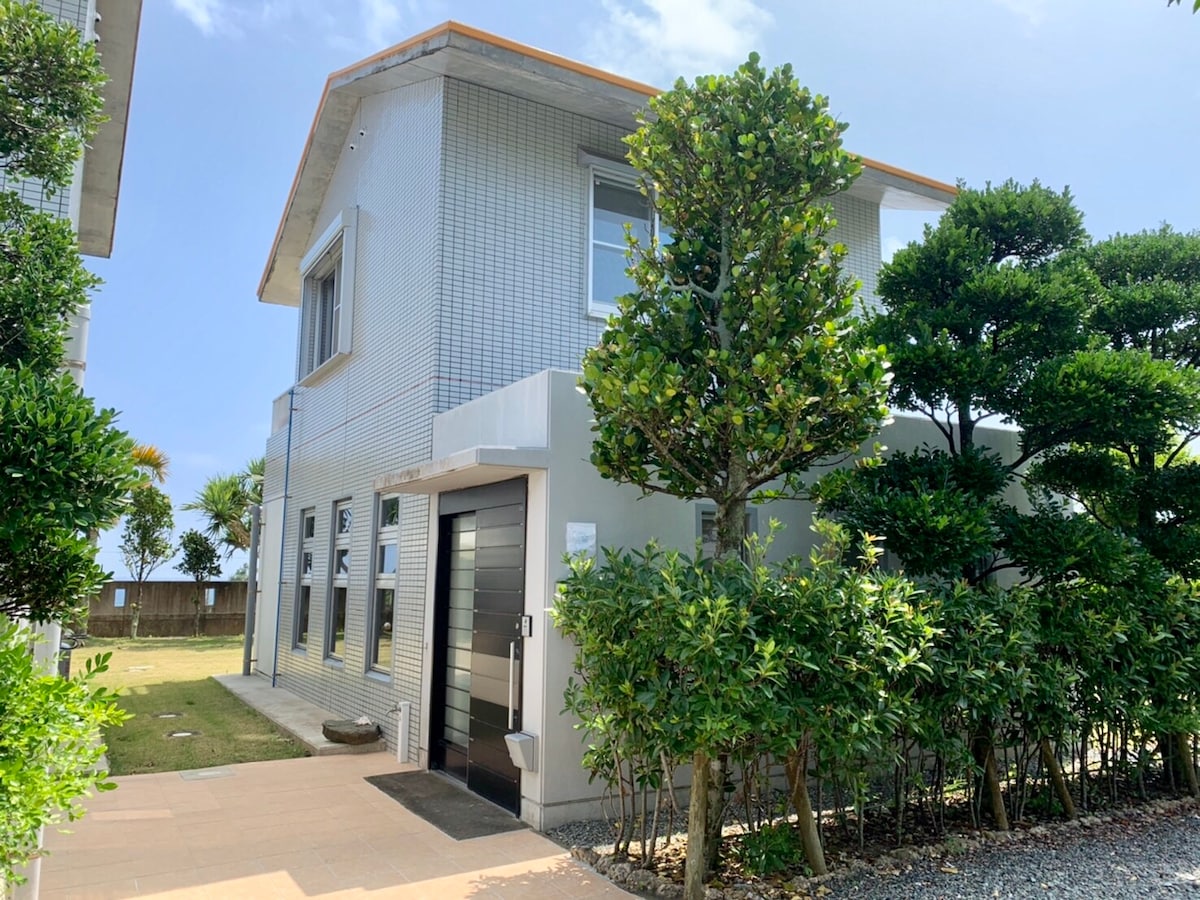 A modern two-story house is presented, featuring a sleek entrance bordered by carefully manicured greenery. The exterior displays a light-colored facade with large windows, allowing natural light to illuminate the interior. A grassy area is visible in the background, contributing to the serene environment.