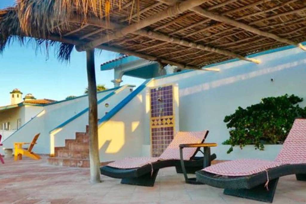 Two lounge chairs with pink patterned cushions are positioned under a thatched roof, offering shade on a spacious patio. A staircase leads to elevated areas, while a plant adds a touch of greenery against the white wall.