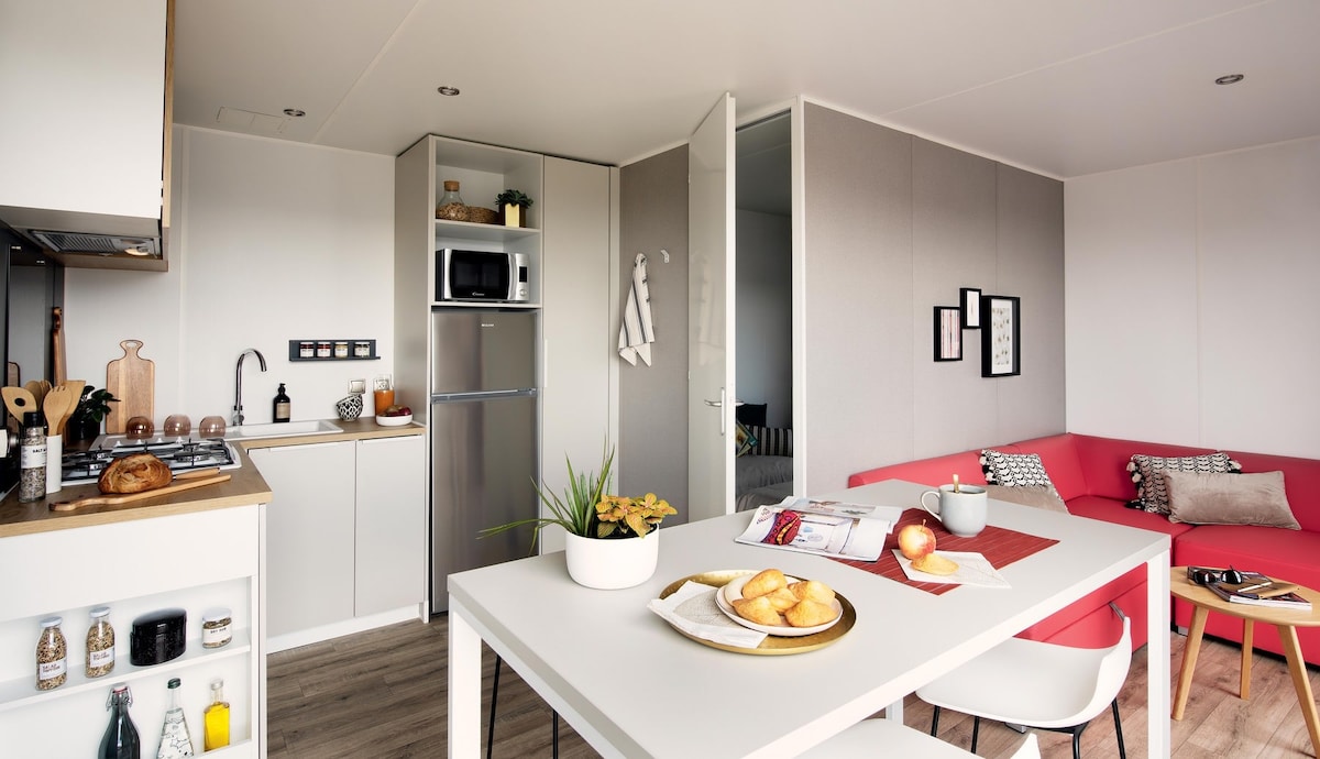 A modern kitchen space features sleek cabinetry and essential appliances, including a stove and microwave. A dining table is set with fresh pastries and fruit. A comfortable red sofa complements the minimalist decor, with natural light illuminating the area.