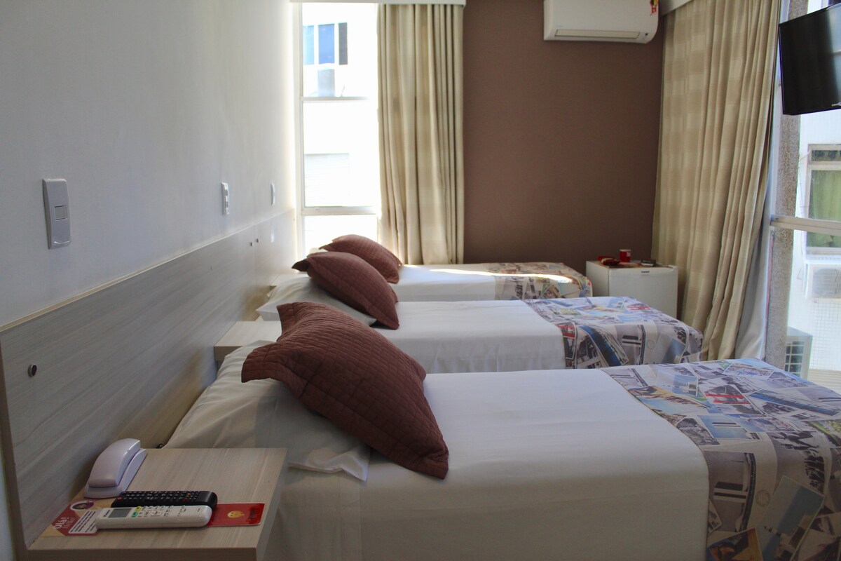 The room features three individual beds with textured pillows, arranged neatly against the wall. A compact work desk is positioned nearby, alongside a mini-fridge. Large windows allow natural light to illuminate the space, complemented by soft, semi-blackout curtains.