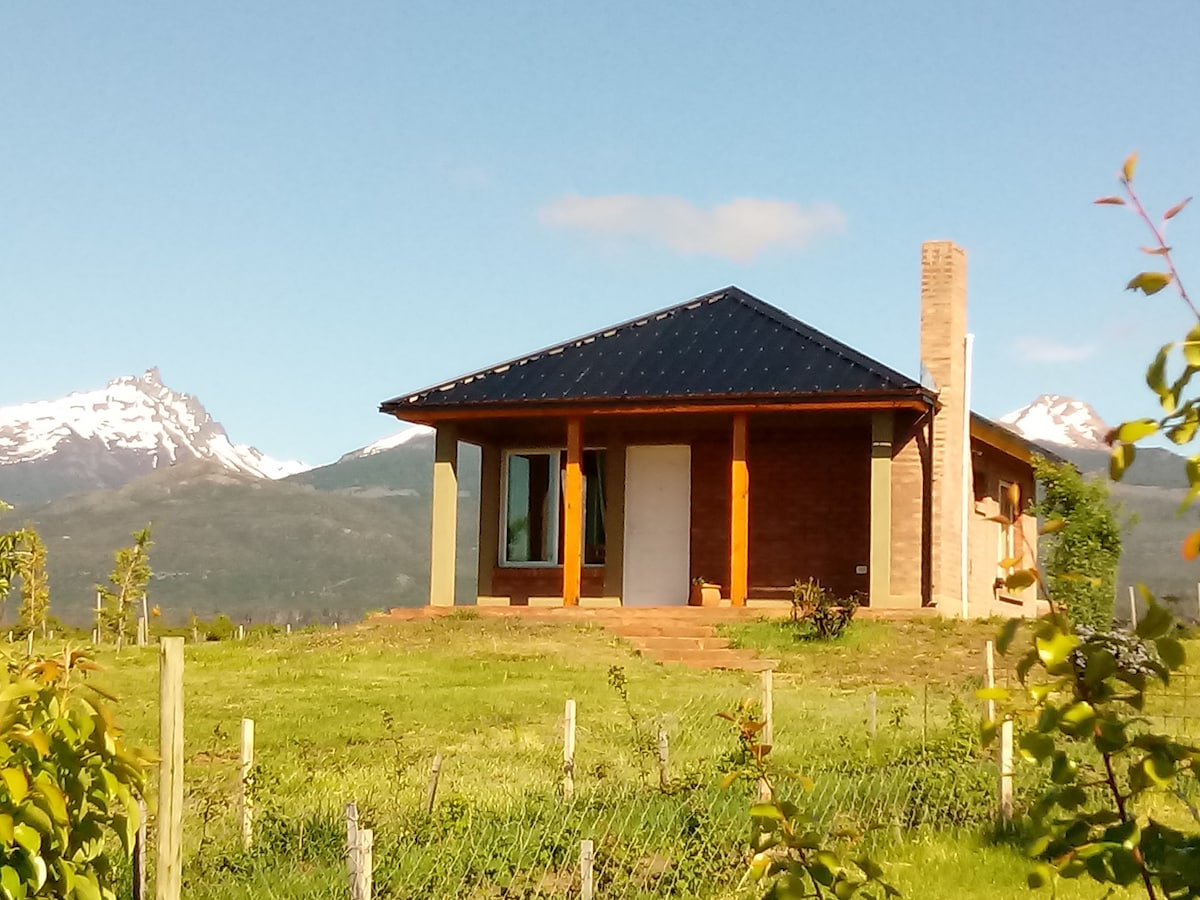 A modern house is set against a backdrop of mountains, featuring a dark roof and large windows. The building's exterior is complemented by brick and wooden accents, while a landscaped area of grass surrounds the structure, showcasing the natural beauty of the location.