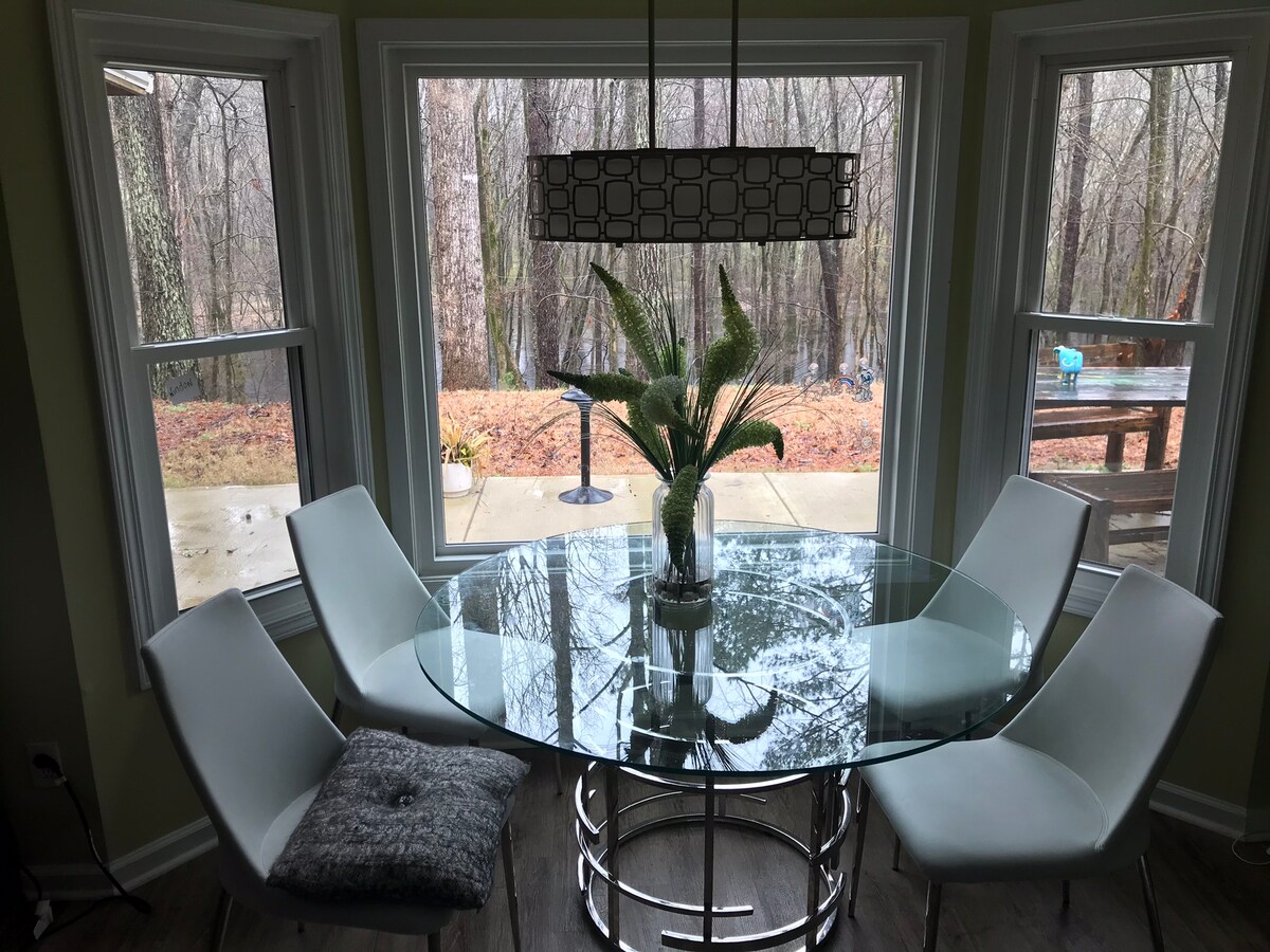 A circular glass dining table is surrounded by four white chairs, providing a modern and airy dining space. Large windows frame a peaceful view of the wooded area outside, letting in soft natural light. A potted plant adds a touch of greenery to the center.