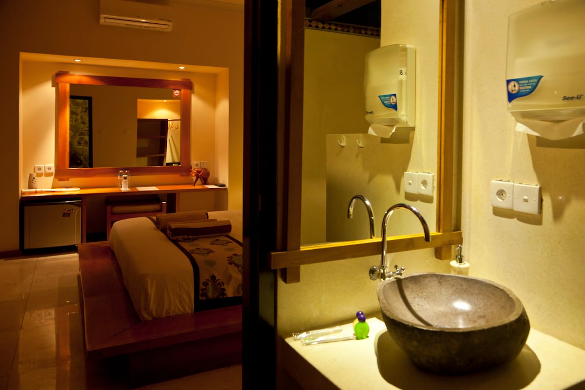 A spacious room is highlighted by soft lighting, featuring a king size bed visible in the background. An outdoor bathroom is presented in the foreground with a stone sink and modern faucet. A mini-fridge can be seen next to the bed area, enhancing convenience.