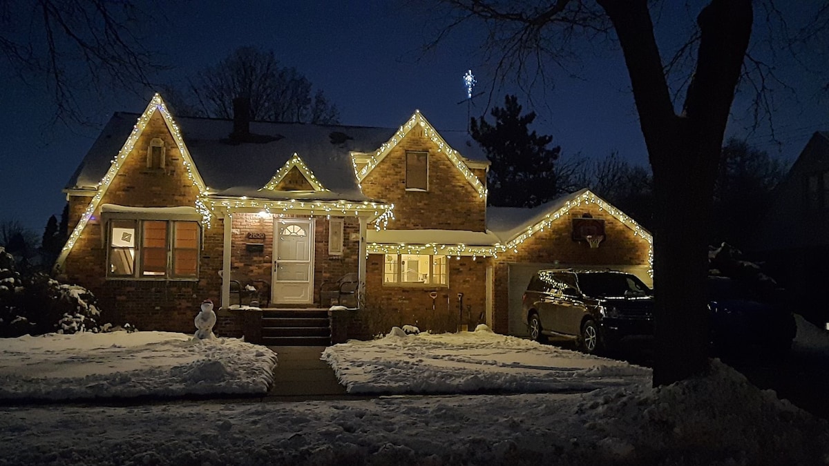 The historic home is adorned with warm white lights, creating a festive atmosphere against the snowy backdrop. The front porch is visible, featuring a welcoming entrance and decorative features. A snowman sits in the front yard, while a vehicle is parked in the driveway.