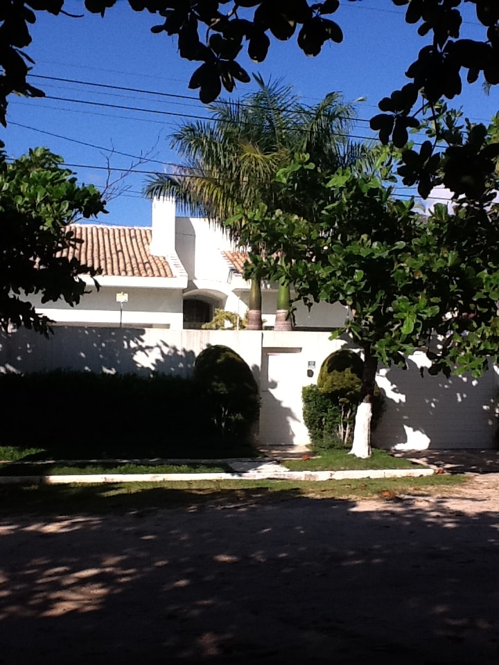 The exterior of the house is framed by lush green foliage, highlighting a welcoming entrance. A palm tree is positioned by the entrance, and the warm sunlight casts gentle shadows on the white wall surrounding the property, contributing to a serene environment.