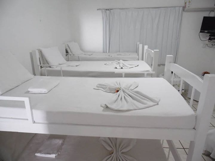 A clean and spacious room contains multiple white bunk beds with crisp white linens. Each bed is adorned with neatly folded towels, and a curtain covers the window, allowing soft light to filter in. A television is mounted on the wall, enhancing comfort.