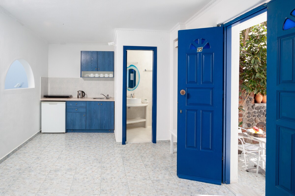 An open living space showcases a kitchenette with blue cabinetry and white appliances. The room features arched windows allowing natural light to illuminate the area. A bathroom is accessible through an archway. Outdoor seating is visible through the blue entrance door.