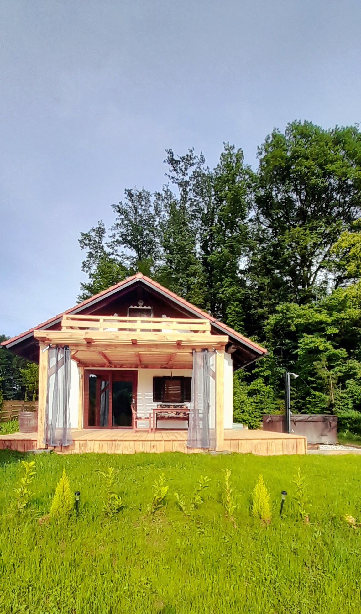A charming cabin is surrounded by lush greenery. The wooden structure features a sloped roof and a covered porch, inviting relaxation. Large windows allow natural light to fill the interior, while blue sky peeks through the trees, enhancing the scenic outdoor view.