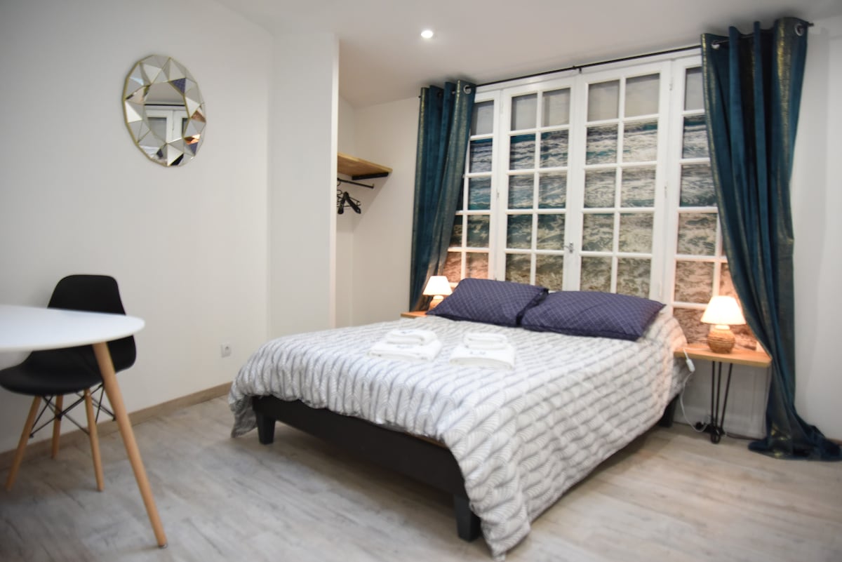 A comfortable sleeping area features a spacious bed with soft bedding and pillows. A large decorative window mural depicting ocean waves is complemented by stylish blue curtains. A round dining table and two chairs provide additional seating, while a mirror and a clothing rack enhance functionality.