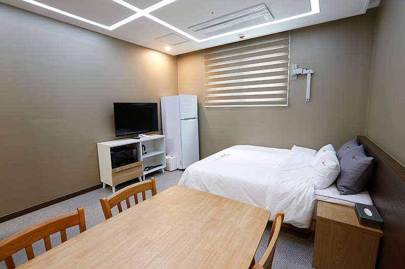A bright room features a spacious bed with neutral bedding positioned against a simple headboard. A 43-inch TV is mounted on the wall opposite the bed, while a refrigerator and microwave are visible next to a wooden dining table with four chairs.
