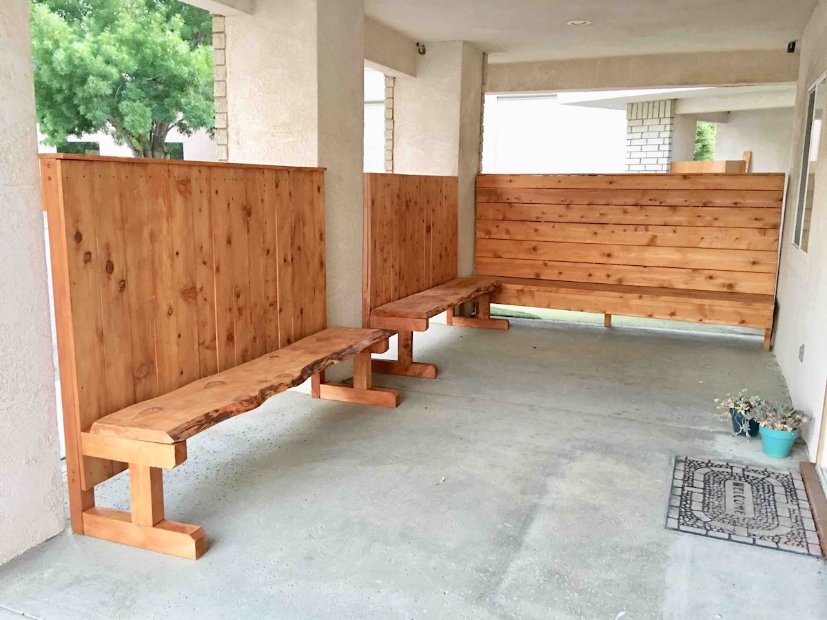 An outdoor seating area is visible, featuring two rustic wooden benches made from natural wood. The benches are positioned against a wooden privacy wall, creating a cozy space for relaxation. Potted plants and a welcome mat add a touch of warmth to the entrance.
