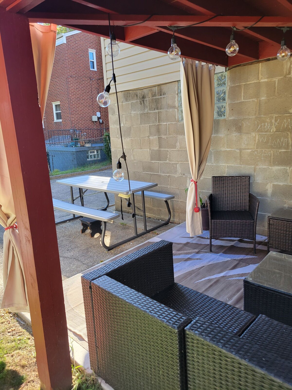 An outdoor space is framed by a red wooden canopy, featuring a rectangular table and chairs made of metal. A seating area with woven furniture provides comfort under soft lighting from hanging bulbs. Privacy curtains drape elegantly, enhancing the tranquil setting.