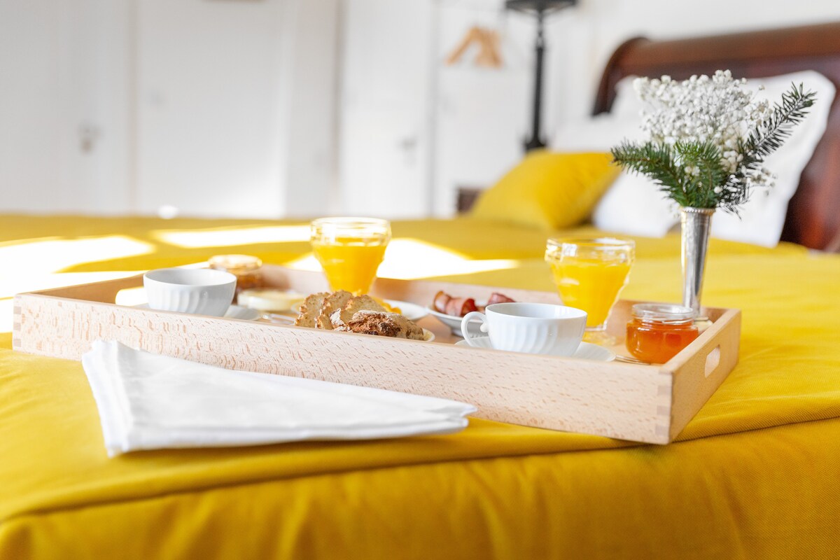 A wooden breakfast tray is placed on a brightly colored bedspread, featuring two small bowls with yogurt, a glass of orange juice, and a sweet dessert. A small vase holds fresh flowers, enhancing the inviting atmosphere of the spacious room.