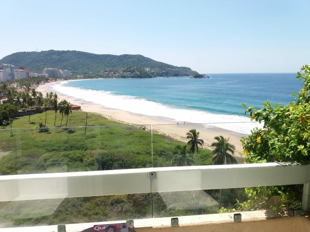 A panoramic view of the beach and ocean is visible from the balcony. Waves gently lap at the sandy shore, while palm trees frame the scene. The landscape includes lush greenery extending toward the beach and distant hills under a clear blue sky.