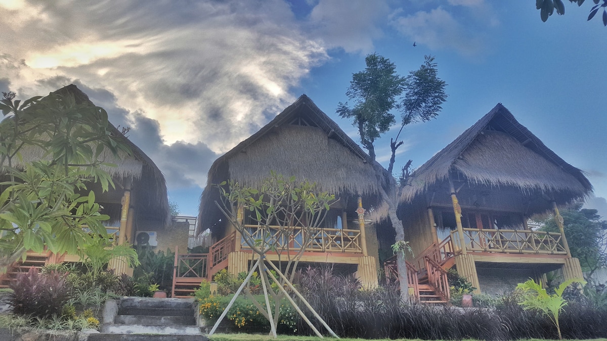 Three bungalows constructed from local bamboo are set against a cloudy sky. Each bungalow features a thatched roof and a porch with wooden railings. Lush greenery and flowering plants surround the bungalows, enhancing the natural setting.
