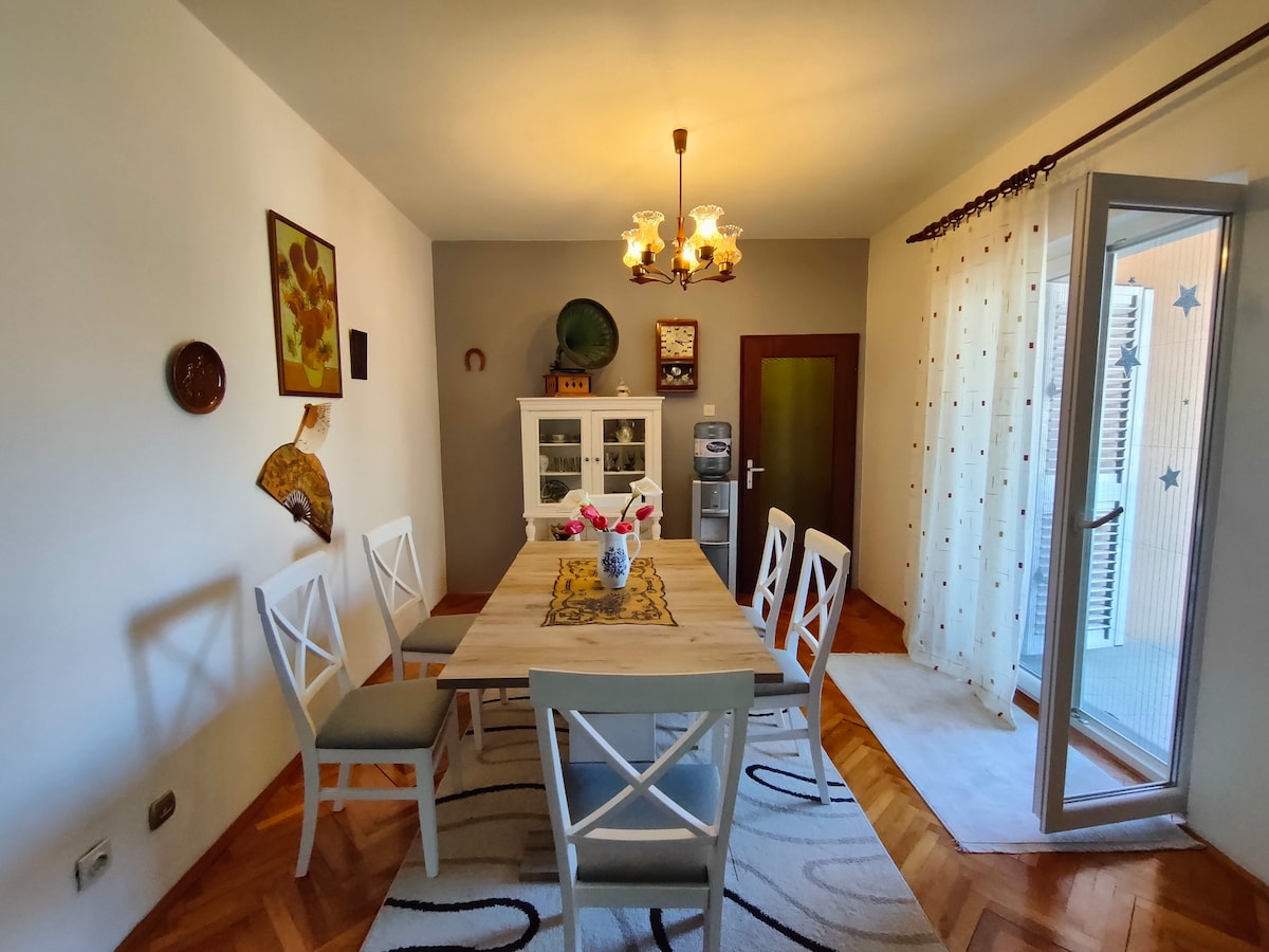 A dining area features a large wooden table surrounded by six white chairs. Natural light enters through the glass doors that lead to the balcony. A rug with a simple pattern lies beneath the table, and a light fixture hangs above, illuminating the space.