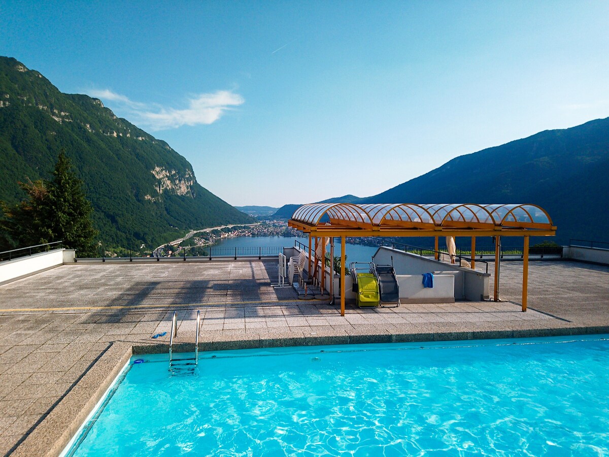 A shared outdoor pool is positioned on a spacious terrace with a vibrant blue water surface. The surrounding mountains rise majestically in the background, with a view of Lake Lugano visible at the far end. Sun loungers and a covered seating area are located nearby.