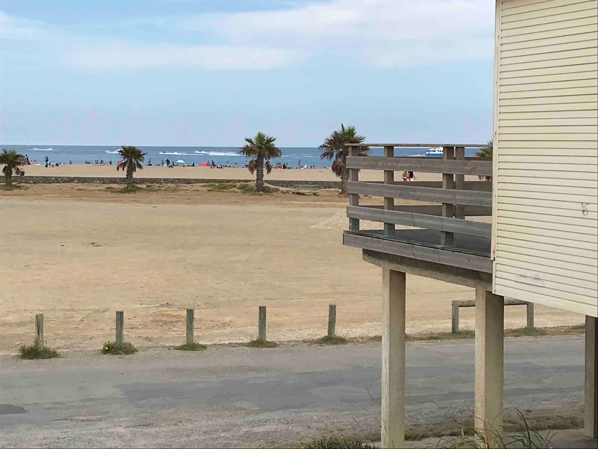 A sandy area is visible, leading to the beach with groups of people enjoying the coastal environment. Palm trees frame the scene, while a wooden deck extends from the adjacent building, providing a potential vantage point for ocean views.