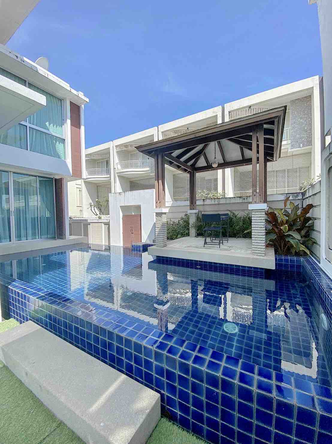 A private mini pool is highlighted in this outdoor space, featuring blue tiles that reflect the sky. A shaded area with a supported roof is visible, alongside lush plants that enhance the setting. Clear, sunny skies contribute to a bright ambiance.