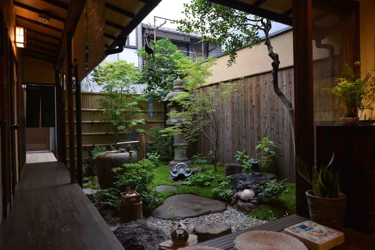 A serene Japanese garden is seen through a wooden-framed entrance, featuring lush greenery, carefully arranged stones, and a traditional stone lantern. The path is lined with smooth stones, and a small water feature adds tranquility to the outdoor space.