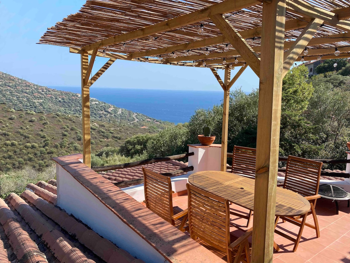 An outdoor terrace features a wooden pergola providing shade, complemented by a round table and four chairs made of natural wood. The backdrop offers a sweeping view of the sea and surrounding hills, highlighting the tranquil natural setting.