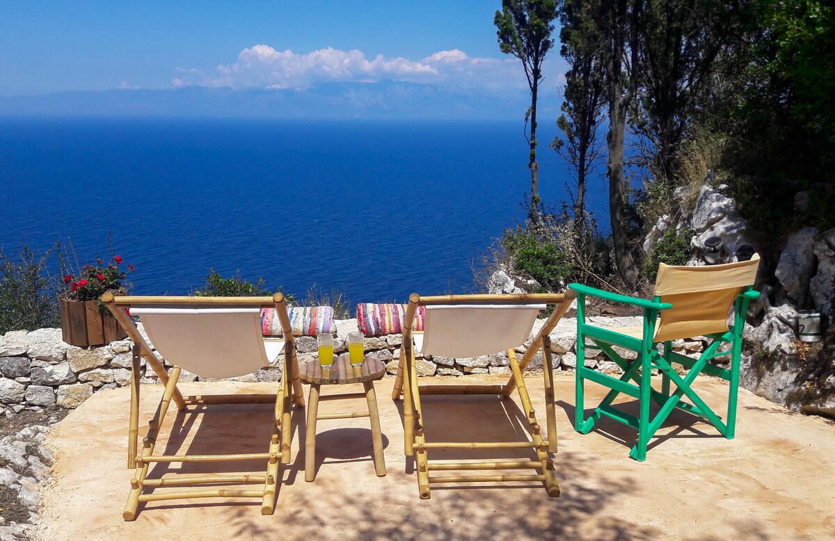 Two wooden lounge chairs are positioned on a stone patio overlooking the sea. A small table is situated between the chairs, holding two beverages. Lush greenery and rocky terrain frame the view, with distant blue waves extending to the horizon under a clear sky.