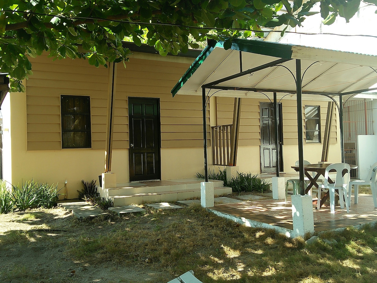 The exterior of the duplex cottage displays a beige facade with dark trim. A covered porch features a small table and white chairs, providing a shaded area. The entrance is marked by a green door, while tall grass and low shrubs frame the property.