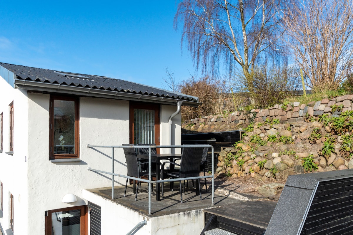 An outdoor terrace is visible, featuring black wicker chairs and a table, surrounded by a stone wall and greenery. The building’s exterior is white, with large windows that allow natural light to enter. A clear blue sky enhances the welcoming atmosphere.
