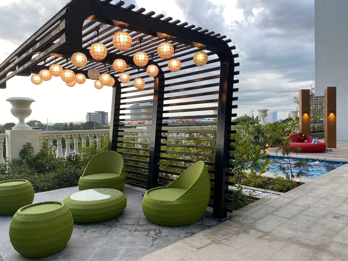 A stylish outdoor seating area features modern green lounge chairs and round ottomans beneath a contemporary pergola. Warm pendant lights are suspended overhead, providing soft illumination. The background includes a glimpse of a tranquil pool and landscaped greenery, offering a relaxing space for guests.