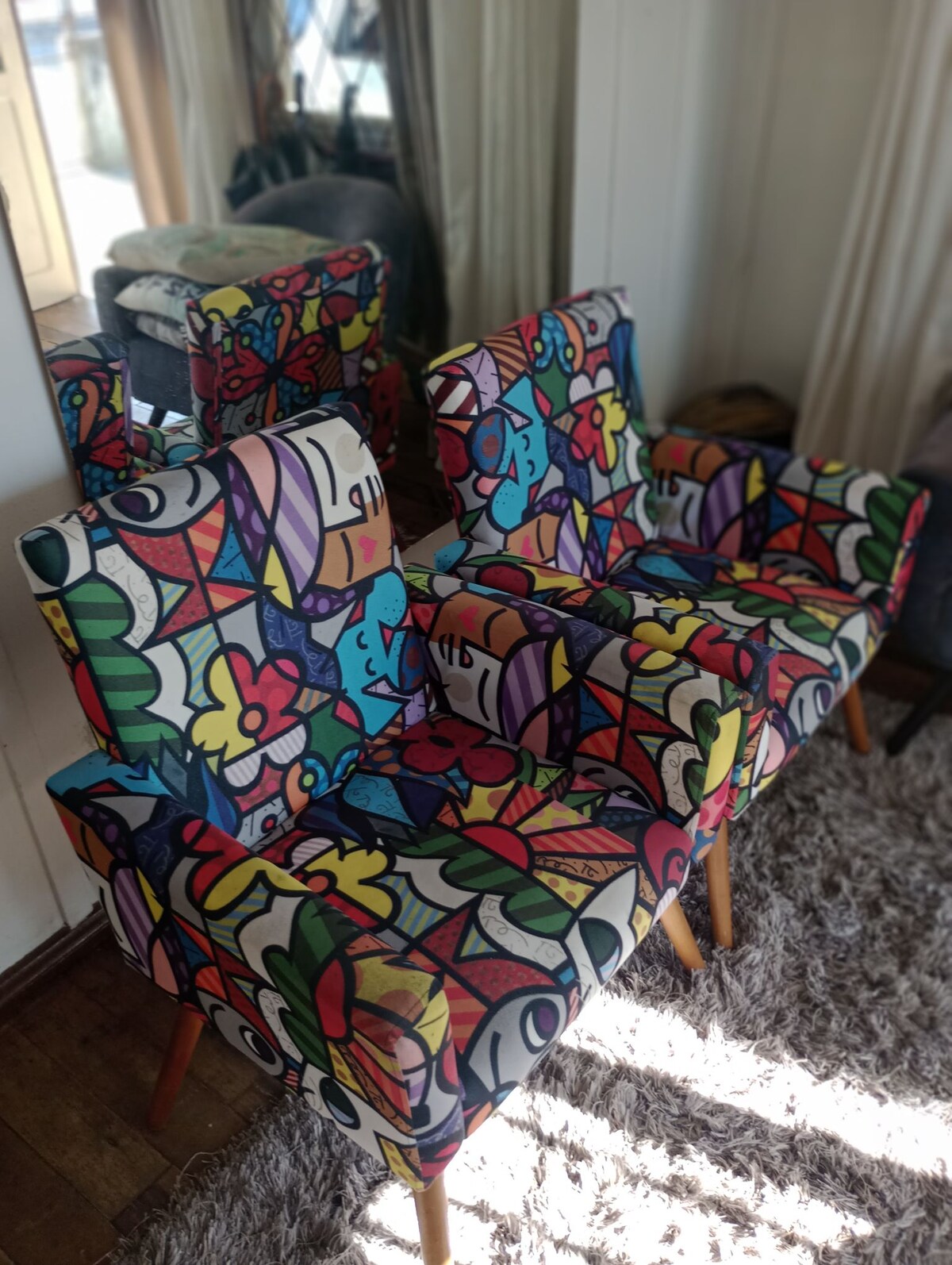 Two armchairs feature vibrant, multicolored upholstery with floral and abstract patterns. They are positioned next to a mirror, which reflects the playful design. The chairs rest on a light-colored rug, enhancing the cozy atmosphere of the living space.