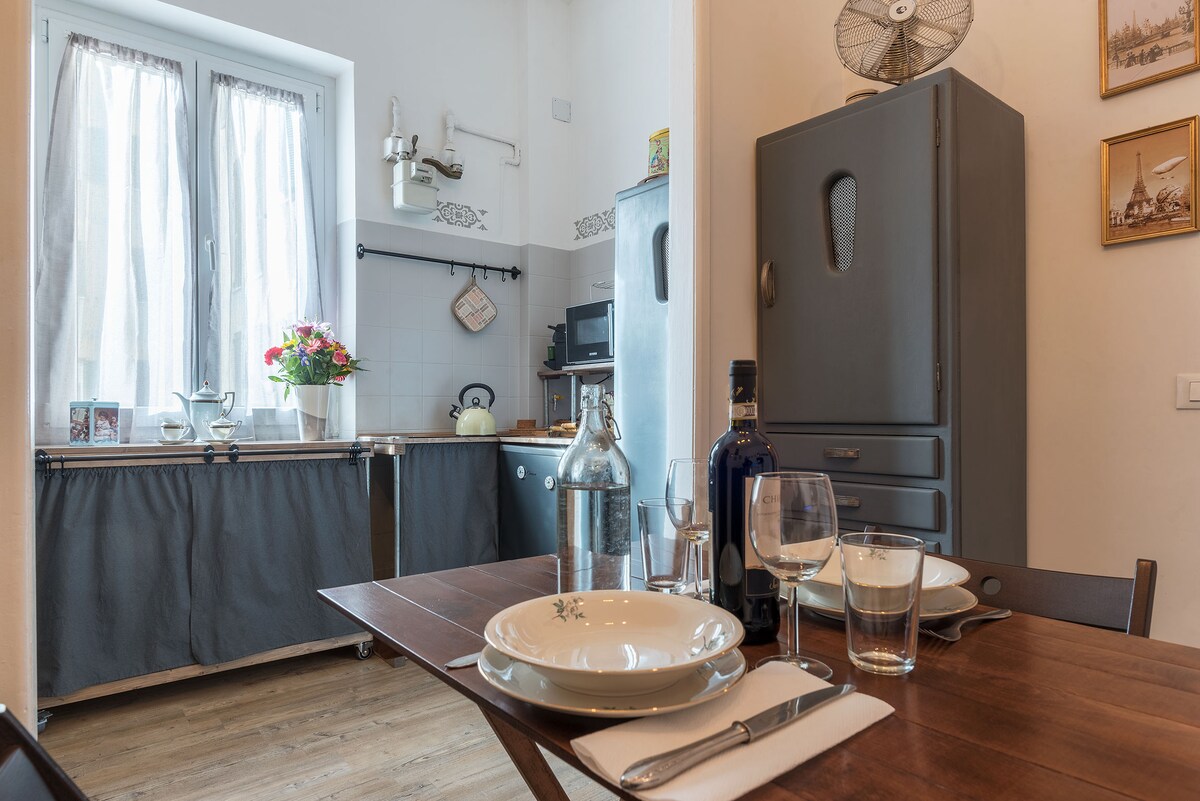 A dining area is set with plates, glasses, and cutlery on a wooden table. A vintage gray refrigerator stands nearby, with a microwave and decorative kitchen items visible. Two windows allow natural light, with light curtains softly filtering the sun.