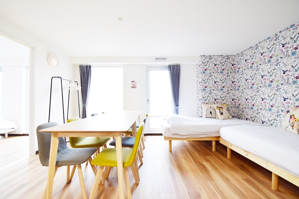 A spacious room is presented with wooden flooring and a light, airy atmosphere. A dining table with six chairs sits centrally, while two beds are positioned against a patterned wall adorned with birds. Natural light filters through large windows with curtains.