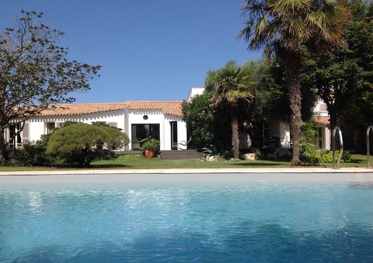 The image showcases a luxury villa set amidst a lush garden, featuring palm trees and various greenery. A clear swimming pool reflects the blue sky, while the villa's white exterior and terracotta roof contrast beautifully against the vibrant surroundings.