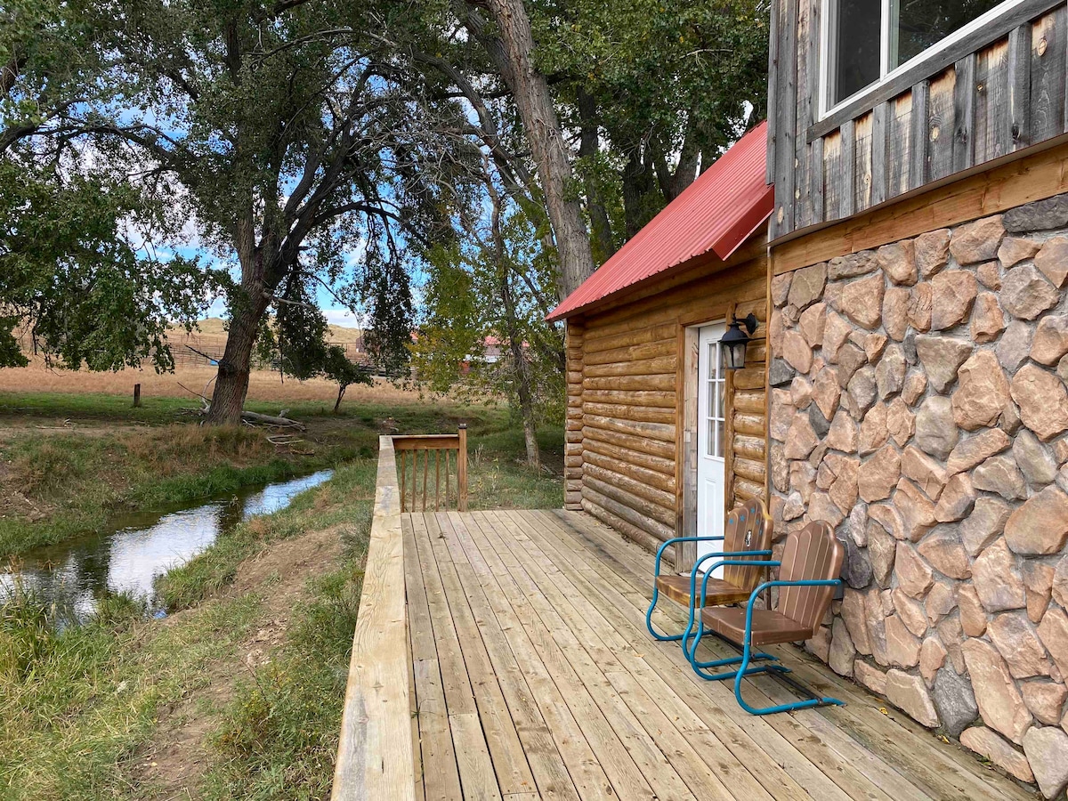 A wooden deck extends from the cozy cabin, featuring two chairs arranged for relaxation. Nearby, a small creek flows gently, framed by trees and open fields, creating a peaceful outdoor setting.