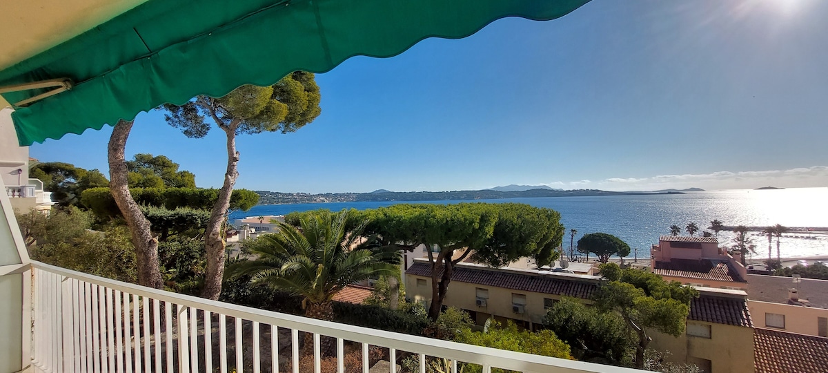 A spacious balcony extends from the apartment, offering panoramic views of the bay and coastline. Lush green trees frame the scene, while the tranquil water reflects the clear blue sky. A nearby sandy beach and distant hills are also visible.