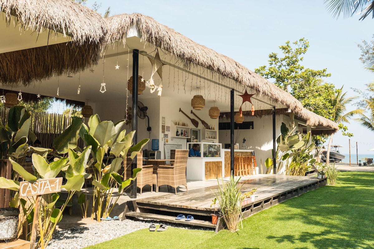 A tropical exterior features a thatched roof covering a wooden deck. Woven chairs and a bar area create a casual outdoor space surrounded by lush greenery. Decorative lights hang gently, offering a relaxed ambiance near the shoreline.