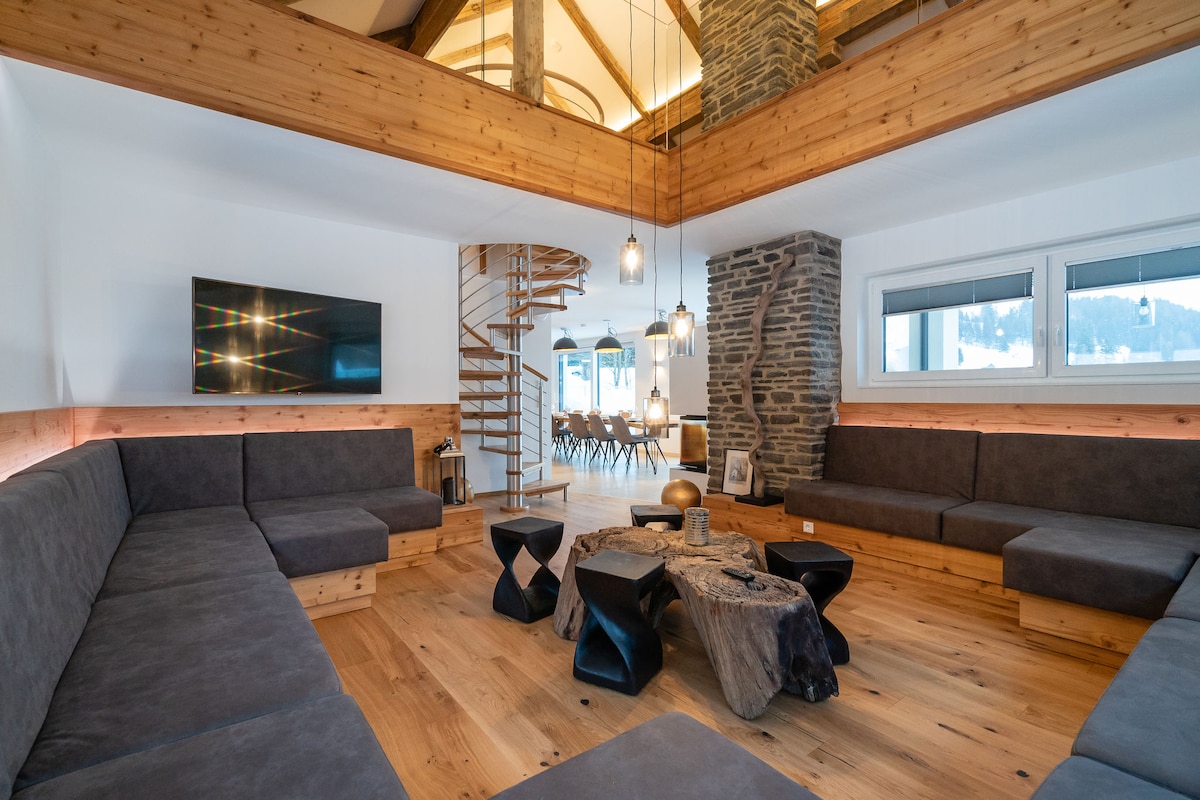 The spacious living room features inviting seating arrangements with dark upholstery around a central wooden table. A stone wall contrasts with the modern décor, while a flat-screen TV is mounted above. Warm lighting fixtures and a spiral staircase add character to the design.