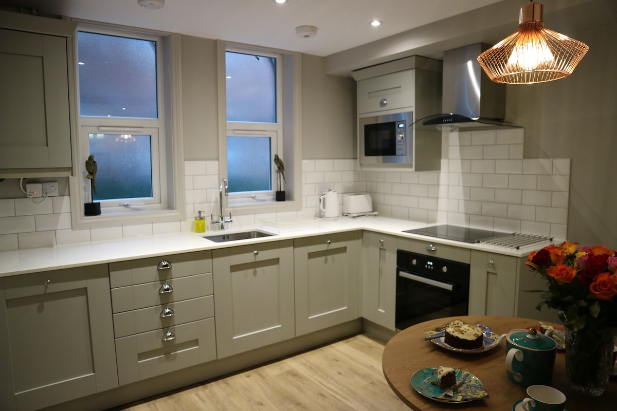A modern kitchen features light grey cabinetry and a white tiled backsplash. Large windows provide natural light. Stainless steel appliances, including a microwave and oven, are integrated into the design. A wooden table is set with a plate of pastries and a vase of roses.
