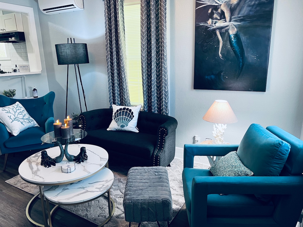 A cozy lounge area features a dark sofa and two accent chairs in a vibrant shade of blue. A contemporary glass table, adorned with decorative candles, rests atop a soft area rug. Artwork depicting a mermaid is displayed on the wall, complementing the room's whimsical theme.