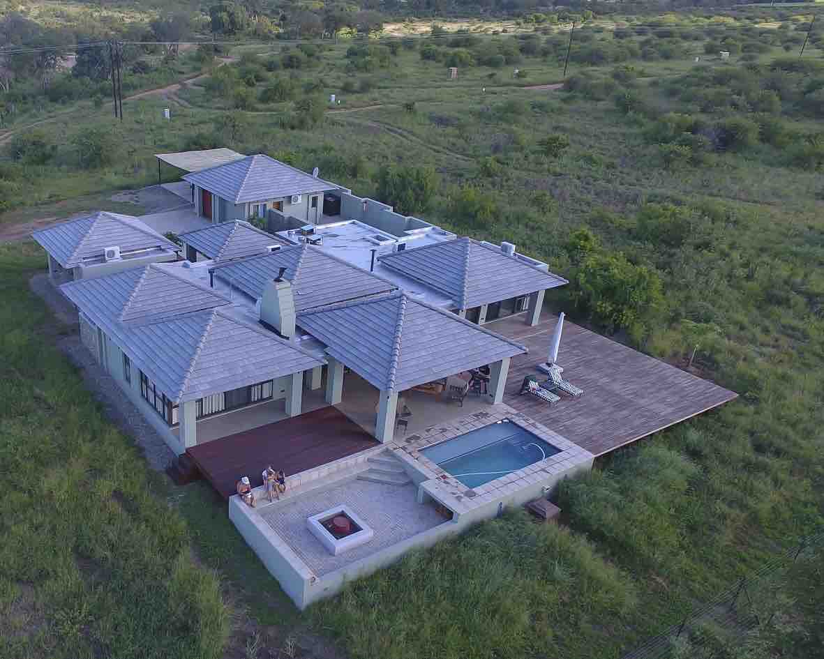 An aerial view captures the spacious lodge featuring a wooden deck, private pool, and outdoor fire pit. The lodge is surrounded by lush greenery and open spaces, with multiple sloped roofs blending harmoniously into the natural landscape.