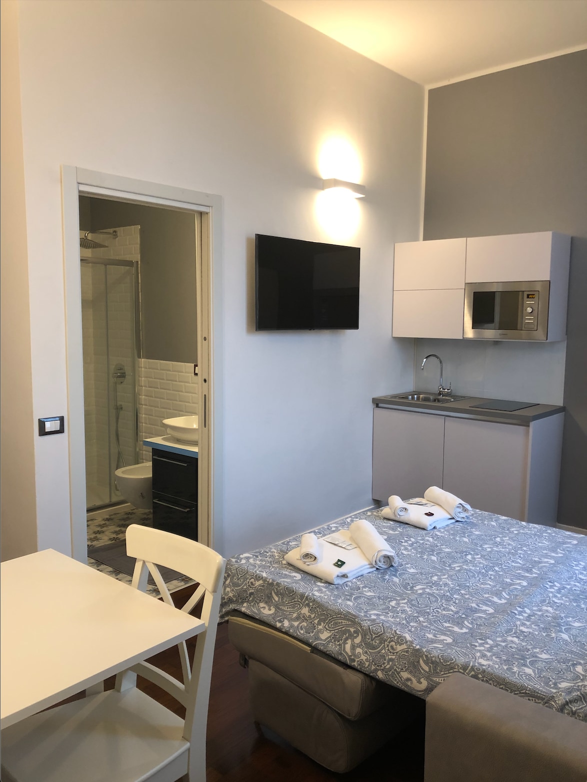 A studio apartment features a cozy sleeping area with a neatly made sofa bed and a patterned blanket. A small dining table with white chairs is positioned nearby. A kitchenette is equipped with modern appliances and a wall-mounted television is mounted above.