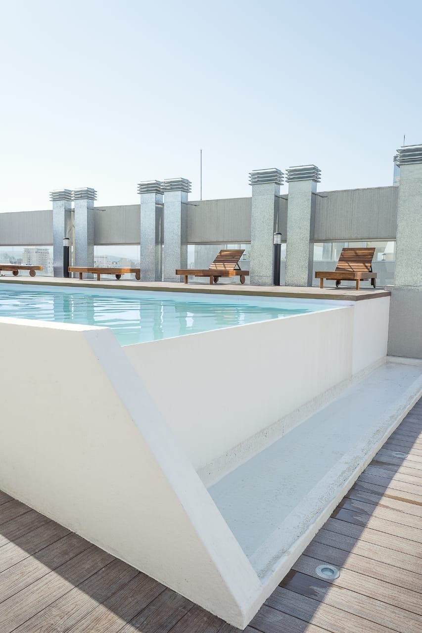 A rooftop pool is surrounded by a wooden deck, featuring sun loungers for relaxation. The minimalist design highlights clean lines and a serene atmosphere, with sunlight reflecting off the water's surface under a clear sky.
