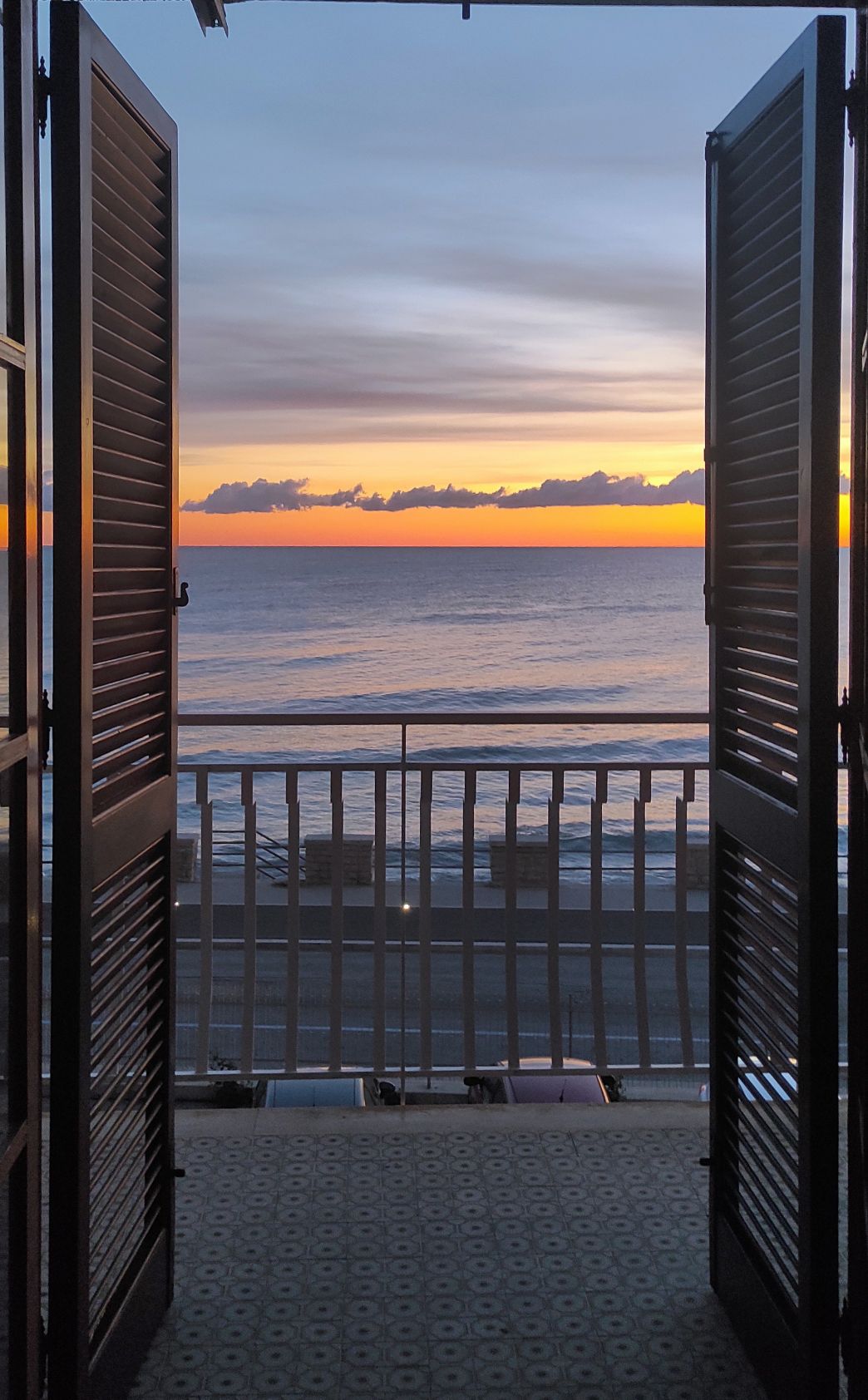 Open louvered shutters frame a serene sunset view over the Adriatic Sea. The vibrant colors of the sky transition from warm orange to soft purple, with gentle waves reflecting the light. A balcony with a white railing is visible, enhancing the coastal ambiance.