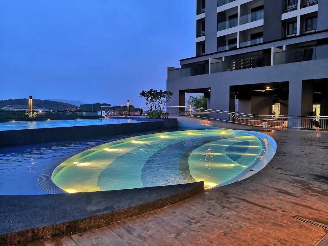 An outdoor swimming pool features a curved design and is illuminated with soft lighting. Surrounding greenery adds a touch of nature. The pool area is adjacent to the building, showcasing a blend of modern architecture and leisure space.