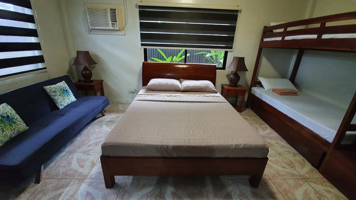 A spacious family room features a queen-sized bed and a double bunk bed, providing ample sleeping options. A folding couch bed offers additional flexibility. Natural light enters through large windows adorned with striped blinds, while two bedside tables with lamps complete the cozy setting.