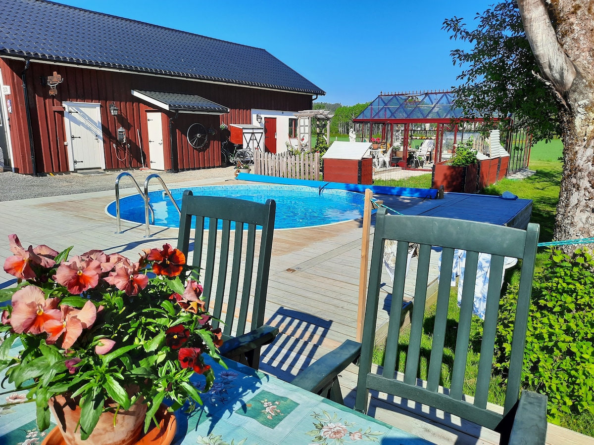 A well-maintained outdoor area features a swimming pool surrounded by a deck. Wooden chairs with a floral arrangement sit at a table, offering a view of the pool and nearby garden structures. Bright sunlight enhances the appeal of the serene environment.