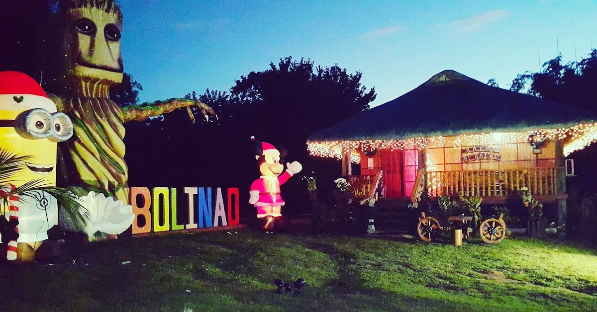 A charming Kubo-style resthouse is set against a twilight sky, surrounded by a grassy area. Colorful decorative lights adorn the exterior. Playful character statues, including a Minion and a Santa Claus, enhance the inviting ambiance, while a bold sign reading 'BOLINAO' adds a cheerful touch.